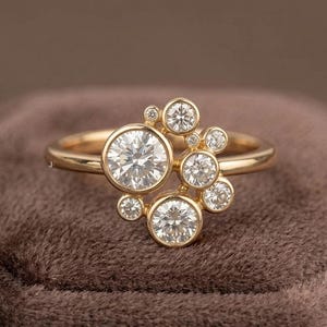 May include: A gold ring featuring a cluster of multiple round, clear, faceted stones set in bezel settings. The stones vary in size and are arranged in an asymmetrical pattern across the top of the polished gold band, creating a cluster design.