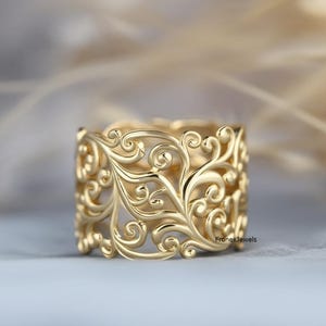 May include: A gold-coloured ring with an intricate openwork design. The ring features a repeating pattern of swirling floral and leaf motifs. The ring is a wide band style, with a polished finish. The ring is likely made of gold.