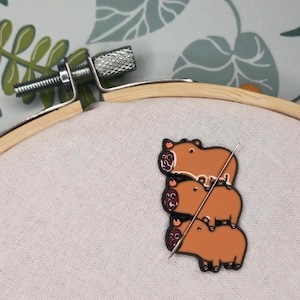 Könnte beinhalten: Eine hellbraune Emaille-Anstecknadel mit drei übereinander gestapelten Capybaras, befestigt auf einem cremefarbenen Stoff, der in einem runden Holzstickrahmen gespannt ist. Die Nadel wird von einer dünnen Metallnadel durch den Stoff gehalten.