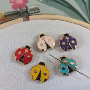 May include: A collection of colorful ladybug-shaped enamel pins with gold accents, each with a unique color scheme: red, pink, purple, yellow, and turquoise. A needle is placed near the pins on a white fabric stretched within an embroidery hoop.