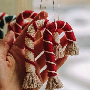 Könnte beinhalten: Zwei rote und weiße gestreifte Garn-Zuckerstangen-Ornamente mit weißen Quasten, die an einer Holzleiste hängen.