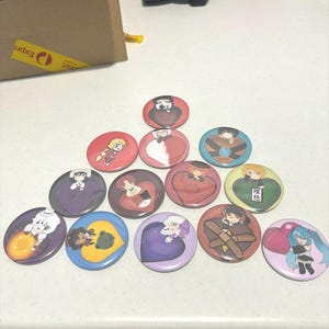 Può includere: Una collezione di 12 spille rotonde con vari disegni colorati che presentano personaggi anime. Le spille sono disposte a forma di triangolo su una superficie bianca.