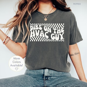 Puede incluir: Una camiseta gris con el texto "Dibs on the HVAC Guy" en blanco con un patrón a cuadros detrás del texto. La modelo lleva la camiseta con unos vaqueros azules.