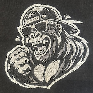 以下が含まれることがあります： 野球帽とサングラスをかけたゴリラの白黒刺繍デザイン。ゴリラは笑顔で、拳を突き上げています。