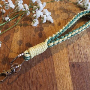 May include: A handmade lanyard in green and cream, with a braided design. It has a silver-coloured clasp and key ring. The lanyard rests on a wooden surface, next to white flowers.