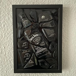 Puede incluir: Una obra de arte de pared con marco negro que presenta piezas rotas de una botella de whisky Jack Daniel's dispuestas sobre un fondo negro. Los fragmentos muestran texto y marca en blanco, incluyendo el logo, 70cl y 40% de alcohol.