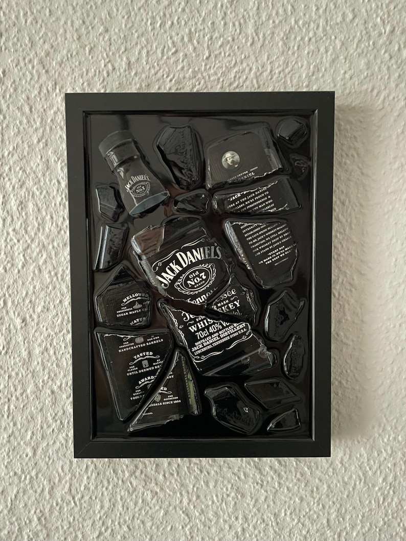 Puede incluir: Una obra de arte de pared con marco negro que presenta piezas rotas de una botella de whisky Jack Daniel's dispuestas sobre un fondo negro. Los fragmentos muestran texto y marca en blanco, incluyendo el logo, 70cl y 40% de alcohol.
