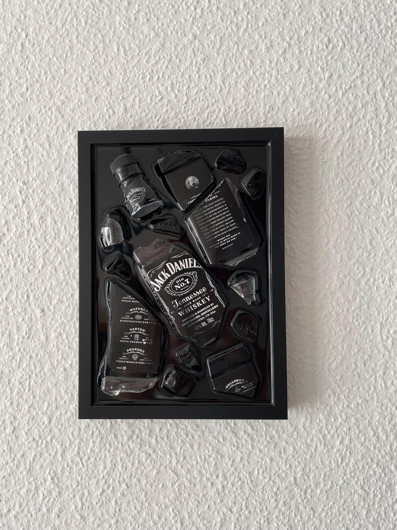 Puede incluir: Un marco rectangular negro tipo caja de sombra que contiene varios fragmentos de vidrio de una botella de whisky Jack Daniel's Tennessee. Las piezas est&aacute;n dispuestas para mostrar las ic&oacute;nicas etiquetas en blanco y negro con el nombre.