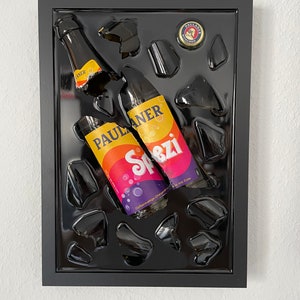 Könnte beinhalten: Ein schwarzes gerahmtes Kunstwerk mit zwei zerbrochenen Glasflaschen von Paulaner Spezi, einem koffeinhaltigen Orangensoda mit Cola, auf schwarzem Hintergrund.