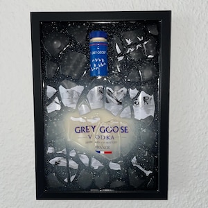 Può includere: Un'opera d'arte incorniciata che presenta una bottiglia in miniatura di Grey Goose Vodka incastonata in un mosaico bianco e nero. La bottiglia è parzialmente immersa in una sostanza bianca, che ricorda la neve o il ghiaccio, con uno sfondo di montagna. L'etichetta recita "GREY GOOSE VODKA DISTILLATA E IMBOTTIGLIATA IN FRANCIA".