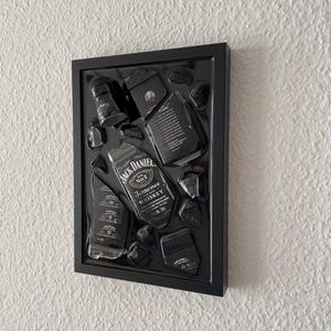 Puede incluir: Un marco negro tipo caja de sombra que muestra una botella de whisky Jack Daniel's Tennessee deconstruida y componentes de la etiqueta. El arreglo incluye la ic&oacute;nica etiqueta en blanco y negro, fragmentos de vidrio y elementos de marca sobre un fondo negro.