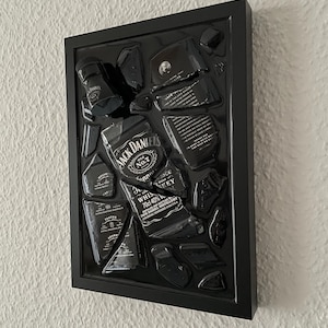Puede incluir: Un marco rectangular negro que contiene una botella de whisky Jack Daniel's rota. Los fragmentos de vidrio est&aacute;n dispuestos para mostrar las ic&oacute;nicas etiquetas en blanco y negro, incluyendo el texto Jack Daniel's Old No. 7 Tennessee Whiskey 70cl 40%.