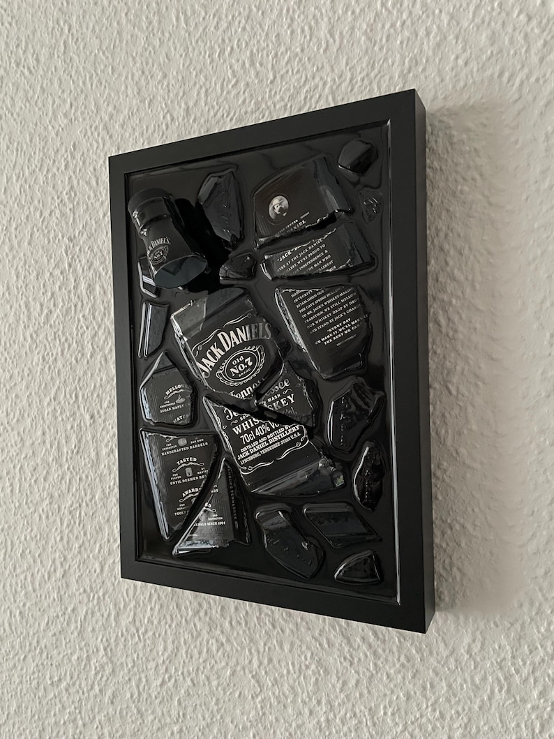 Puede incluir: Un marco rectangular negro que contiene una botella de whisky Jack Daniel's rota. Los fragmentos de vidrio est&aacute;n dispuestos para mostrar las ic&oacute;nicas etiquetas en blanco y negro, incluyendo el texto Jack Daniel's Old No. 7 Tennessee Whiskey 70cl 40%.