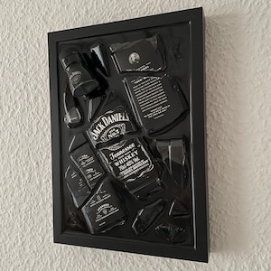 Puede incluir: Una pieza de arte de pared con marco negro que presenta una botella de whisky Jack Daniel's Tennessee rota. Los fragmentos de vidrio est&aacute;n dispuestos dentro del marco, mostrando las ic&oacute;nicas etiquetas en blanco y negro con texto como Jack Daniel's Old No. 7.