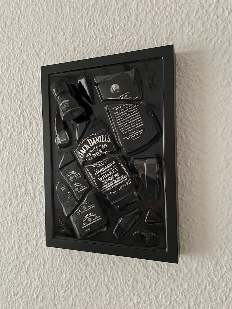 Puede incluir: Una pieza de arte de pared con marco negro que presenta una botella de whisky Jack Daniel's Tennessee rota. Los fragmentos de vidrio est&aacute;n dispuestos dentro del marco, mostrando las ic&oacute;nicas etiquetas en blanco y negro con texto como Jack Daniel's Old No. 7.
