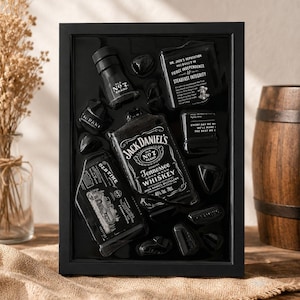 Puede incluir: Un marco de exhibici&oacute;n negro que contiene una botella de whisky Jack Daniel's Tennessee, una botella m&aacute;s peque&ntilde;a y dos placas rectangulares con texto blanco, sobre un fondo negro con varias piedras decorativas negras dispersas.