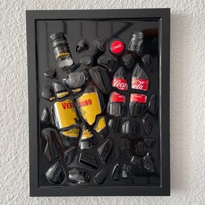 Könnte beinhalten: Ein gerahmtes Kunstwerk mit Fragmenten von Flaschen und Dosen, darunter Coca-Cola und eine gelbe Flasche mit dem Text "VETERANO". Die Teile sind auf einem schwarzen Hintergrund angeordnet, mit schwarzen, glänzenden, unregelmäßigen Formen.