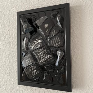Puede incluir: Una pieza de arte mural con marco negro que presenta una botella de whisky Jack Daniel's Tennessee deconstruida. La exhibici&oacute;n incluye varios fragmentos de vidrio y etiquetas dispuestos dentro de una caja negra, mostrando la marca y el texto.