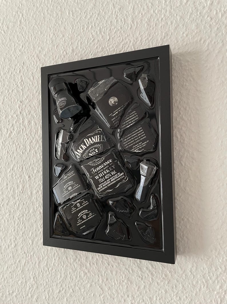 Puede incluir: Una pieza de arte mural con marco negro que presenta una botella de whisky Jack Daniel's Tennessee deconstruida. La exhibici&oacute;n incluye varios fragmentos de vidrio y etiquetas dispuestos dentro de una caja negra, mostrando la marca y el texto.
