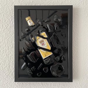 Pode incluir: Uma obra de arte emoldurada em preto que apresenta uma garrafa de Rothmann's Weinbrand, um brandy alemão, incrustado em resina preta. A garrafa está quebrada em pedaços e espalhada pela superfície da resina.