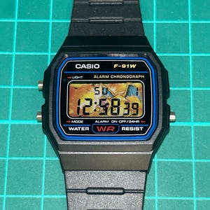 May include: Black Casio F-91W digital watch with a rectangular face and black strap. The watch face displays the time, with the text "CASIO F-91W" at the top. The watch has a blue border and the time displayed is 12:58:39.