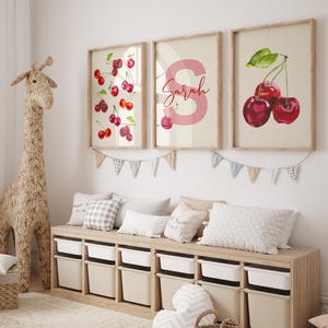 Könnte beinhalten: Drei gerahmte Kunstdrucke mit Kirschillustrationen und einem rosa Buchstaben mit dem Namen "Sarah". Die Kunst hängt über einem Holzaufbewahrungsschrank mit weißen Behältern. Eine gewebte Giraffe schmückt den Raum.