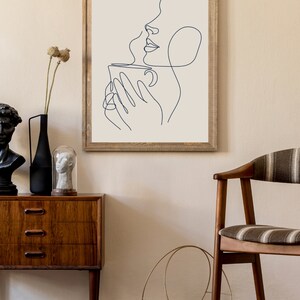May include: A framed line art print of a person holding a cup of coffee. The print is in a wooden frame and is hanging on a beige wall.
