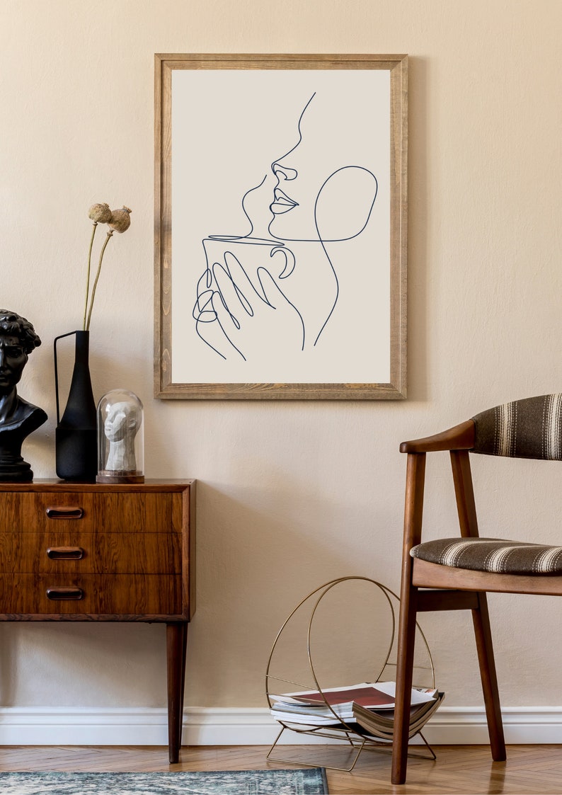 May include: A framed line art print of a person holding a cup of coffee. The print is in a wooden frame and is hanging on a beige wall.