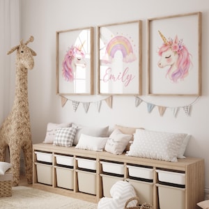 May include: Three framed wall art prints featuring watercolour illustrations of unicorns with pink manes and floral crowns, and a central rainbow with the name Emily in cursive script, displayed above a wooden storage bench with white bins.
