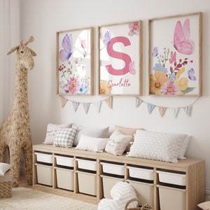May include: Three framed prints with floral and butterfly designs. The center print features a pink letter "S" and the name "Scarlette". The prints are hung on a white wall above a wooden shelf with storage bins.