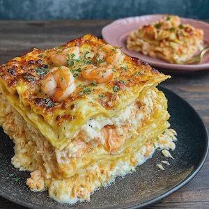 May include: A layered lasagna dish with prawns and creamy white sauce, served on a black plate. The top layer is golden brown, with prawns and green herbs. Another plate with a portion of lasagna is in the background.