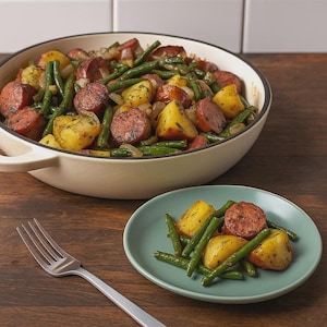 May include: A hearty meal of sausage, potatoes, and green beans in a cream-colored pan and a teal plate. The sausage is sliced and browned, the potatoes are golden, and the green beans are bright green. A silver fork rests on the wooden table.