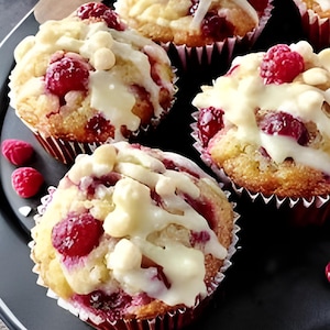May include: Close-up of several muffins with fresh raspberries and white chocolate chips. The muffins are in red and white paper cases and drizzled with white icing. The baked goods are on a dark grey plate.