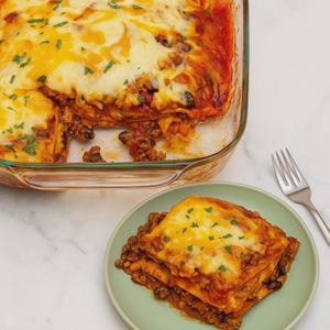 May include: A baked casserole in a clear glass dish, with a portion served on a green plate. The dish features layers of pasta, meat, black beans, and melted cheese, garnished with chopped herbs. A silver fork rests beside the plate.