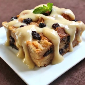 May include: A square slice of bread pudding topped with a creamy white sauce and dark raisins. A sprig of fresh mint is on top of the pudding.