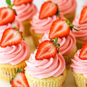 May include: Close-up of cupcakes with pink frosting and strawberry halves. The cupcakes have a yellow sponge base and are arranged on a white surface. A delicious treat.