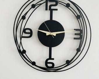 May include: A circular wall clock featuring a black metal frame with overlapping rings and small circular accents. The clock face has large black numerals at the 12, 3, 6, and 9 o'clock positions, a solid black center, and thin gold-colored clock hands.