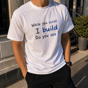 Peut inclure: Un t-shirt blanc à manches courtes avec un texte en noir et bleu qui indique : While you scroll .. I build Do you see ? Le vêtement présente une encolure ronde et une coupe standard, offrant un aspect épuré pour une tenue décontractée.