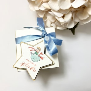 Può includere: Una scatola regalo bianca legata con un nastro di raso azzurro. Un'etichetta a forma di stella con bordo dorato presenta un'illustrazione di un personaggio e il testo "ABBI CURA DI SPLENDERE" in corsivo rosso. Un bouquet di fiori bianchi è sullo sfondo.