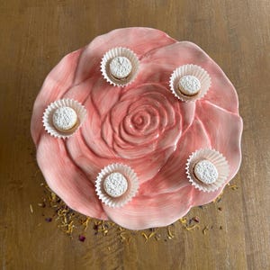 Könnte beinhalten: Ein rosa, rosenförmiger Keramik-Servierteller enthält fünf kleine, runde Gebäckstücke in weißen Papierförmchen. Die Backwaren sind mit Puderzucker bestäubt. Der Teller steht auf einer Holzoberfläche.