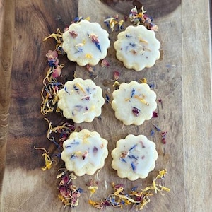 May include: Six round sugar cookies decorated with white icing and colorful edible flowers on a wooden cutting board.