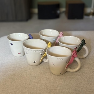 May include: Five off-white ceramic mugs with small painted motifs and colourful ribbon bows attached to the handles. One mug features the text "GOOD MORNING" and another features the text "MAMA". The mugs have various small painted designs including flowers, hearts, and insects.