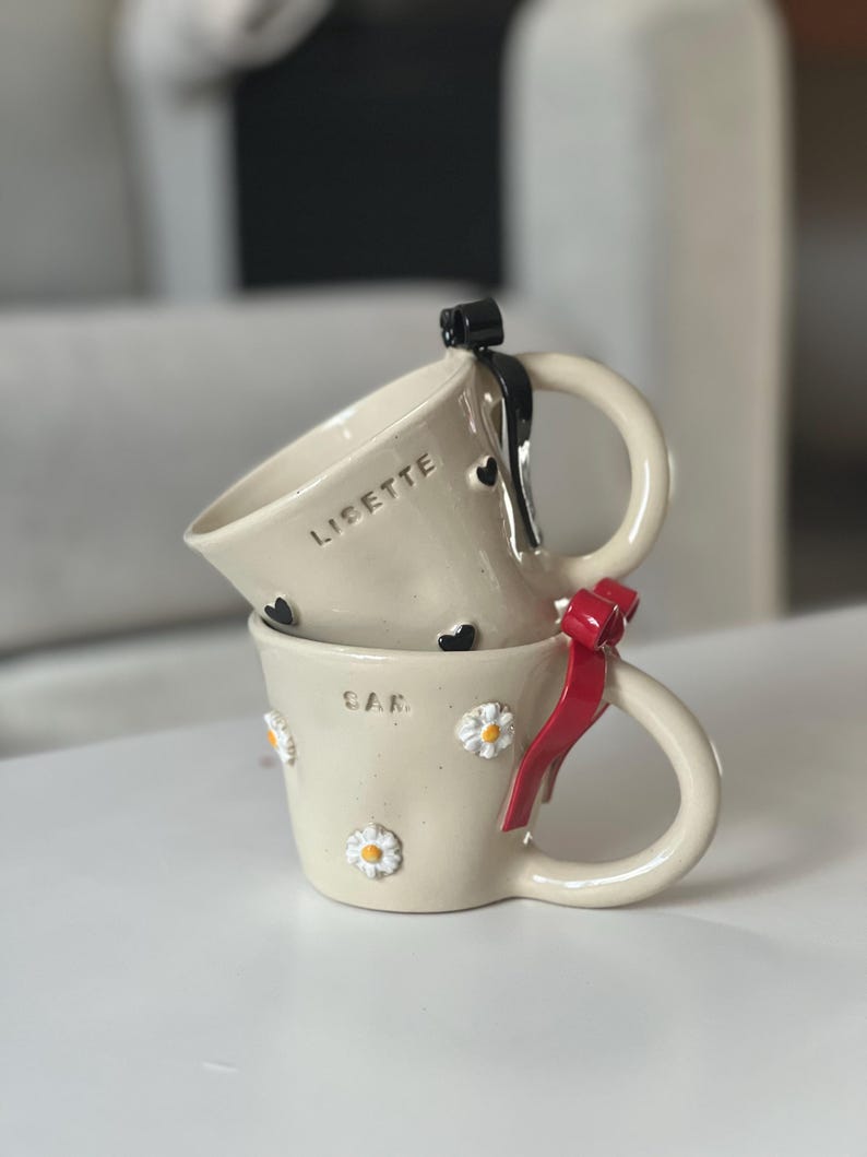 May include: Two stacked ceramic mugs. The top mug has the name "LISETTE" and black heart accents. The bottom mug has the name "SAR" and daisy accents. One mug has a red bow, and the other has a black bow.