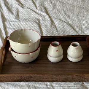 May include: Two stacked cream-coloured ceramic bowls with scalloped edges and small red and pink heart patterns, alongside two matching cream-coloured ceramic bud vases with narrow openings, all featuring red and pink heart motifs.