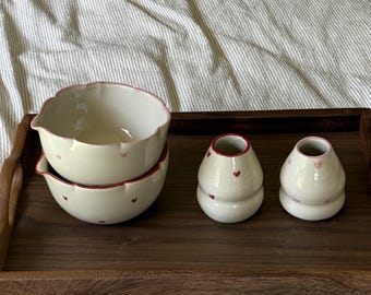 May include: Two stacked cream-colored ceramic bowls with scalloped edges and small red and pink heart patterns, alongside two matching cream-colored ceramic bud vases with narrow openings, all featuring red and pink heart motifs.
