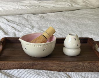 May include: A ceramic matcha bowl with a light pink interior and cream exterior featuring small painted flowers and the text "LIVE LAUGH MATCHA" in black, accompanied by a bamboo whisk and a matching cream-colored ceramic whisk holder.