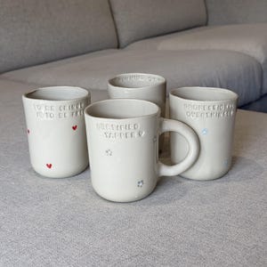 May include: Four off-white ceramic mugs with rounded shapes. One mug features a handle and the text "CERTIFIED YAPPER" with small flower icons. Another reads "TO BE CRINGE IS TO BE FREE" with red hearts, and a third says "PROFESSIONAL OVERTHINKER".