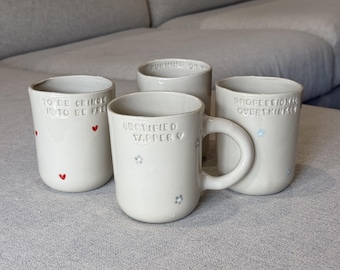 May include: Four off-white ceramic mugs with rounded shapes. One mug features a handle and the text "CERTIFIED YAPPER" with small flower icons. Another reads "TO BE CRINGE IS TO BE FREE" with red hearts, and a third says "PROFESSIONAL OVERTHINKER".