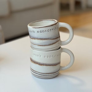 May include: Two cream-coloured ceramic mugs, stacked, with brown and grey horizontal stripes. The top mug reads "INDY'S GODMOTHER", the bottom mug reads "INDY'S GODFATHER". Both mugs have a rounded handle.