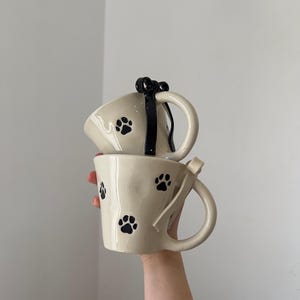 May include: Two cream-colored ceramic mugs with black paw prints. One mug has a black bow detail on the handle, and the other has a cream-colored ribbon detail. The mugs are stacked on top of each other.